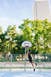 Back view of full body african american sportsman jumping and throwing ball in basketball hoop on sports ground in sunny day