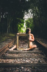 Full length of naked woman with mirror sitting on railroad track at forest