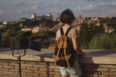 Rear view of mid adult man with backpack standing by retaining wall against cityscape