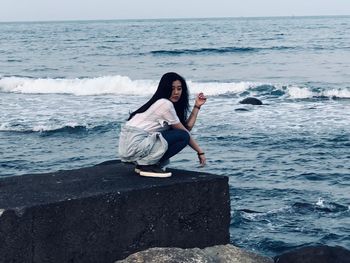 Full length of young woman crouching on concrete wall by sea