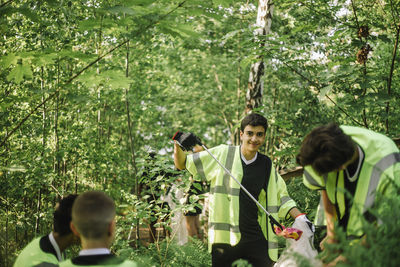 Portrait of smiling teenage boy picking plastic garbage amidst green plants