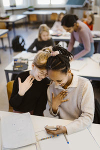 High angle view of schoolgirls gesturing while using smart phone sitting at desk in classroom