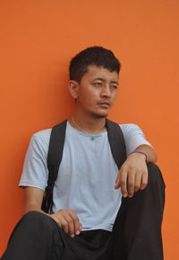 An asian guy looking sideways, sitting against orange wall background with copy space 