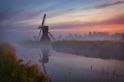 Traditional windmill by lake against sky during sunset