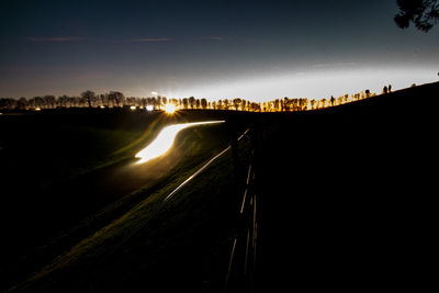 Illuminated road by silhouette city against sky at night