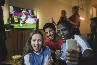 Fans taking selfie with mobile phone while friends watching soccer match on tv