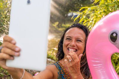 Beautiful smiling middle aged woman taking a selfie sitting on a pink flamingo shaped inflatable toy