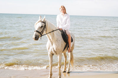 Young red haired woman in white shirt riding white horse on seascape background