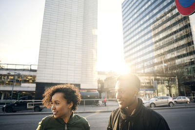 Teenage girl and boy looking away while standing on roadside against buildings