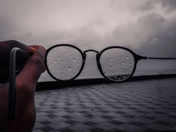 Close-up of hand holding wet eyeglasses against sky