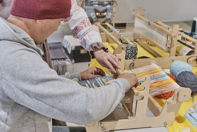 Senior asian man weaving small rug with pattern on manual table loom, at masterclass on weaving.