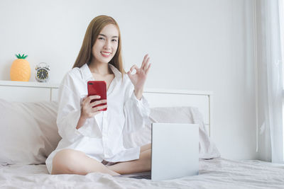 Portrait of cheerful young woman using phone with laptop on bed at home