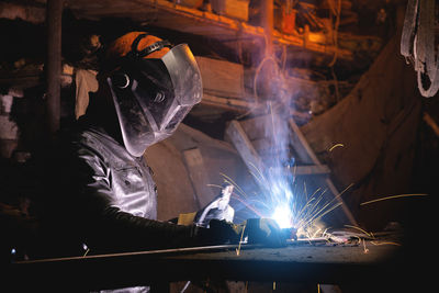 A male welder in a leather jacket carries out welding work in a home shed against the background of