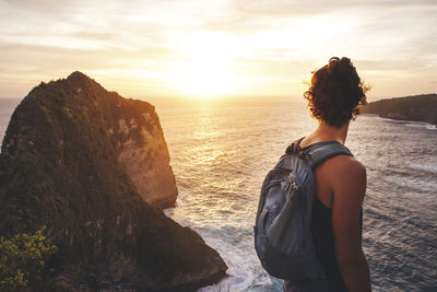 Side view of man with backpack standing on mountain over sea against sky during sunset