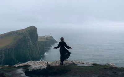 Woman in a black dress standing on rock by sea against sky, neist point scotland 