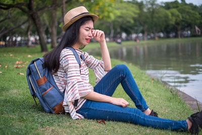 Side view of worried woman with backpack sitting by lake in park