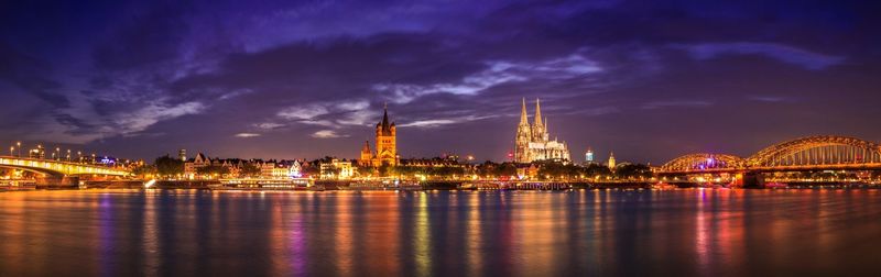 Illuminated buildings by river against cloudy sky