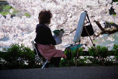 Side view of woman painting while sitting against flowering tree