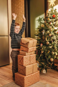 Cheerful boy with arms raised standing by christmas presents at home