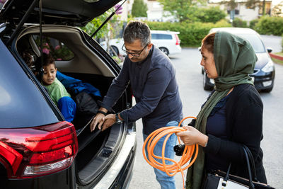 Mother holding pipe while father keeping electric charger by son in car