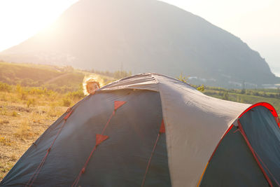 Cheerful child looks out from behind a tourist tent in the mountains with a panoramic view at dawn. 