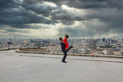 Man carrying woman while standing on terrace against cloudy sky