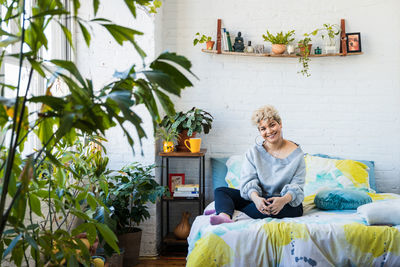 Smiling woman sitting on bed against wall in bedroom at home