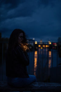 Woman sitting with illuminated string lights on railing against river at night