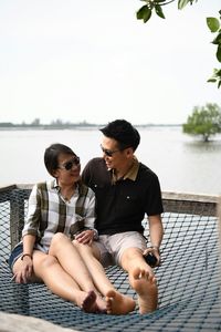 Couple sitting on net over lake against clear sky