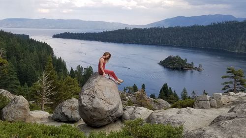 Young woman sitting on rock by lake against sky