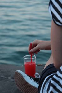 Midsection of woman with drink sitting on jetty at sea