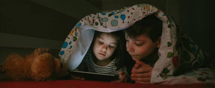 Siblings using digital tablet on bed in darkroom