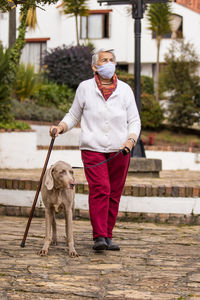 Full length of senior wearing mask with dog walking outdoors