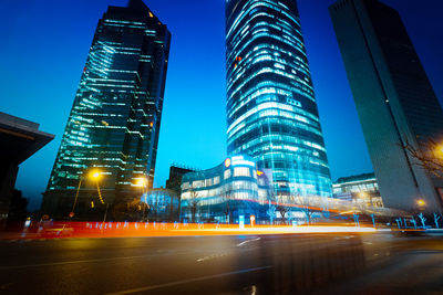 Light trails on city street by buildings against sky at night