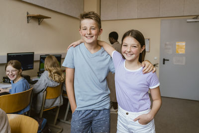 Portrait of smiling male and female friends standing with arms around in computer class at school