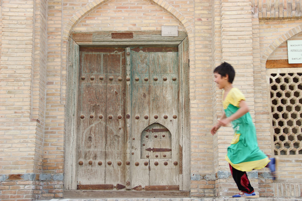 one person, full length, standing, child, side view, childhood, casual clothing, architecture, females, day, entrance, brick, women, door, building exterior, girls, hairstyle, leisure activity, fashion, outdoors, innocence