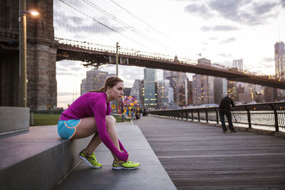 Woman tying shoe lace while sitting on steps against brooklyn bridge