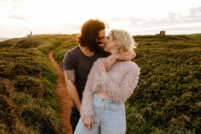 Bearded man embracing and kissing blond girlfriend while standing on grassy cliff at sunset in aviles, spain