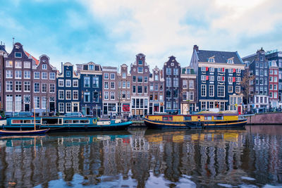 Buildings in city by river against cloudy sky