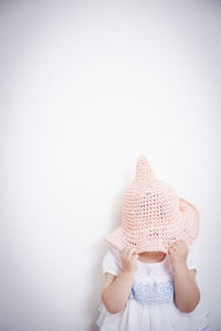 Girl covering face with knit hat while standing against wall at home
