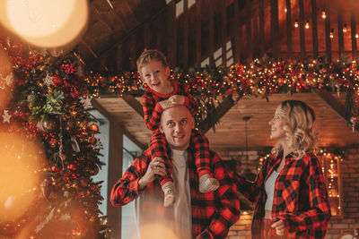 Smile active parents with small son in red checkered sleepwears waiting santa indoor.