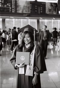 Portrait of smiling woman in graduation gown holding diploma while standing in college