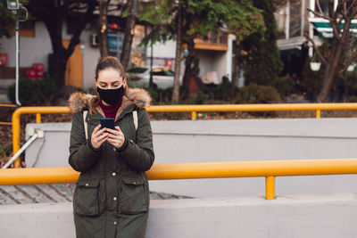Young woman wearing mask using smart phone while standing outdoors