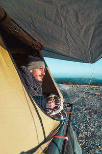 Side view of adult camper lying in tent and enjoying landscape of hilly valley at sunrise during trekking adventure