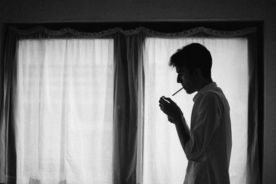 Side view of young man smoking while standing against window at home