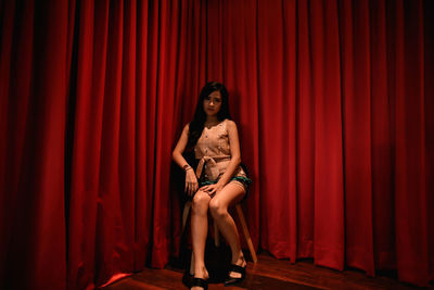 Portrait of young woman sitting on stool against curtain