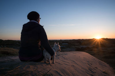 Rear view of woman with dog sitting on landscape at sunset