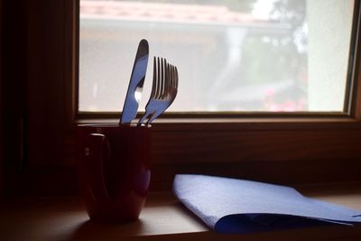 Close-up of coffee cup with fork on table against glass window at home