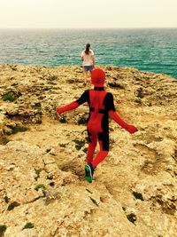 Rear view of boy in costume with mother on rock formation by sea