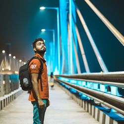 Side view of young man standing on footbridge in city at night